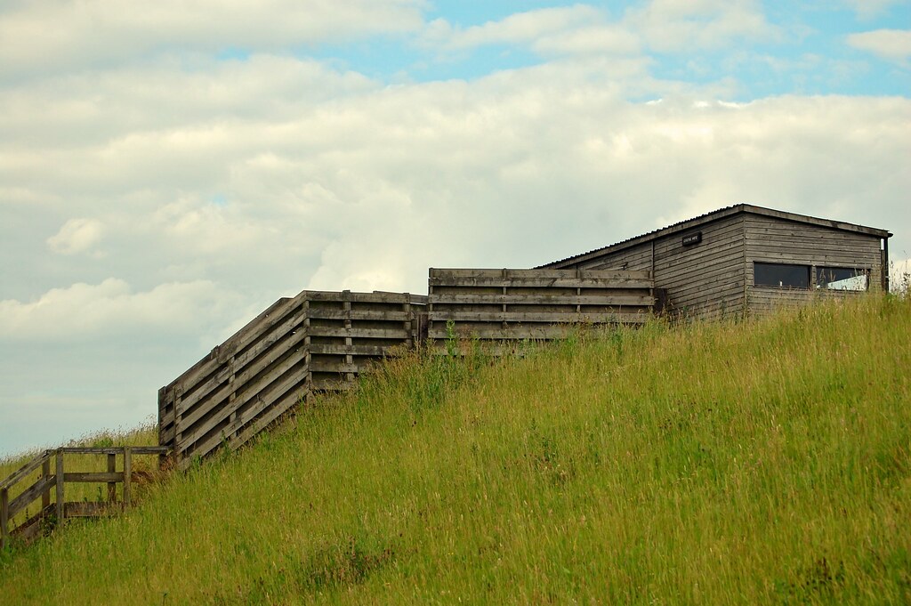 Wooden structure with a window sits on a grassy hill under a cloudy sky.