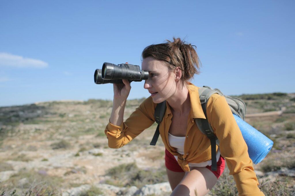 A woman hiker observes the landscape through binoculars on a sunny day outdoors.