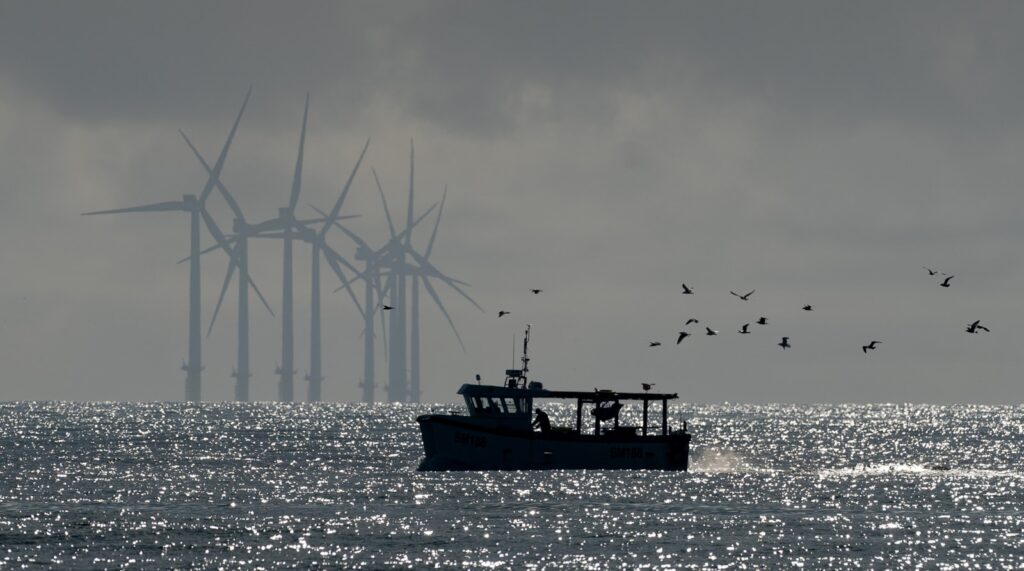 Dark silhouettes of birds fly above a boat on the water, with wind turbines visible in the distance.