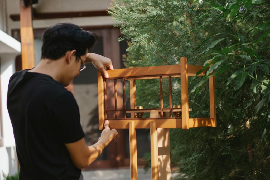 Back view of concentrated young male joiner installing wooden handmade bird table in yard near trees.