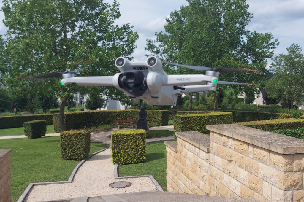 A white DJI Mini 3 Pro drone hovers over a manicured green space.