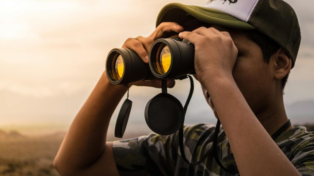 A young man wearing a cap uses binoculars to observe the scenic landscape outdoors during daylight.