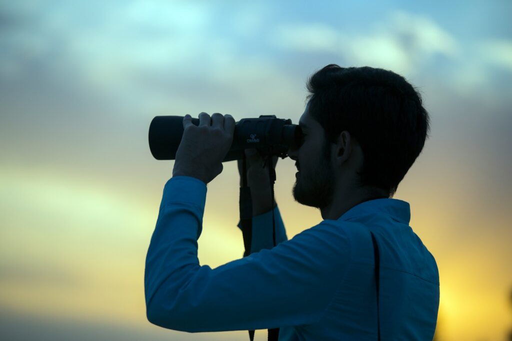 A silhouetted man in a blue shirt looks through black binoculars against a muted sky.
