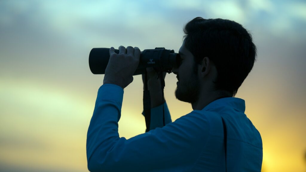 A silhouetted man in a blue shirt looks through black binoculars against a muted sky.