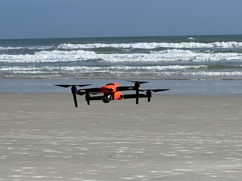 An orange Autel EVO II drone flies over a sandy beach near the ocean.