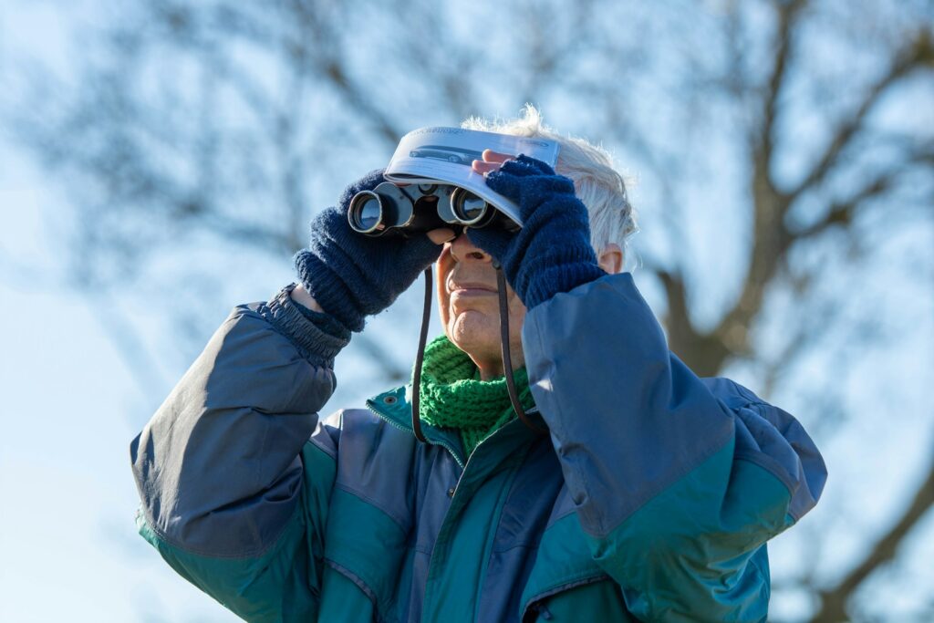 A person wearing a blue and green jacket, gloves, and a scarf uses binoculars.