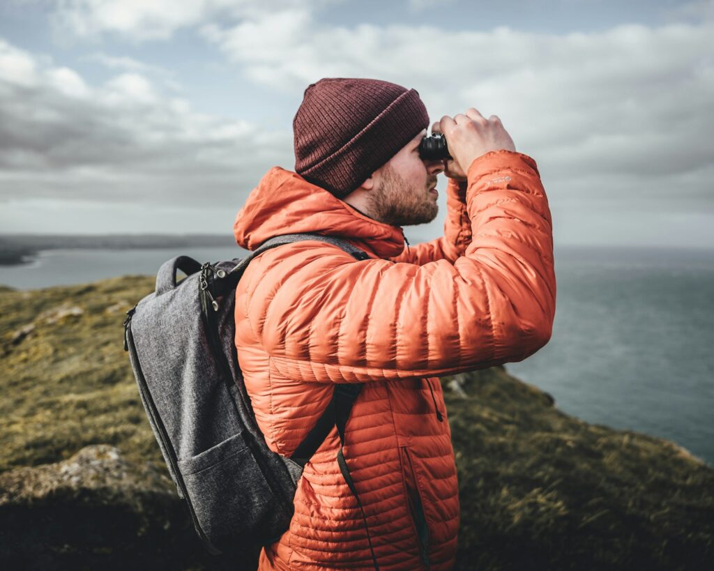A person wearing a maroon beanie, orange jacket, and backpack uses binoculars outdoors.
