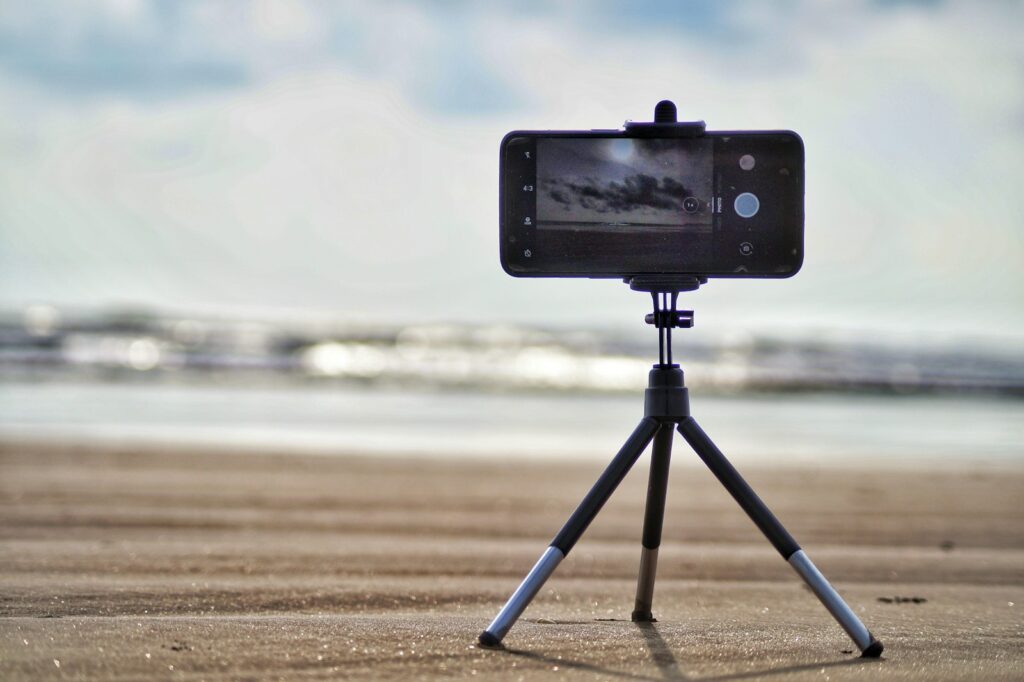 A smartphone is mounted on a small tripod on a sandy surface.