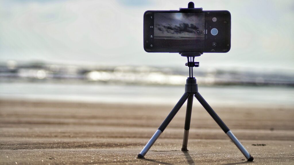 A smartphone is mounted on a small tripod on a sandy surface.