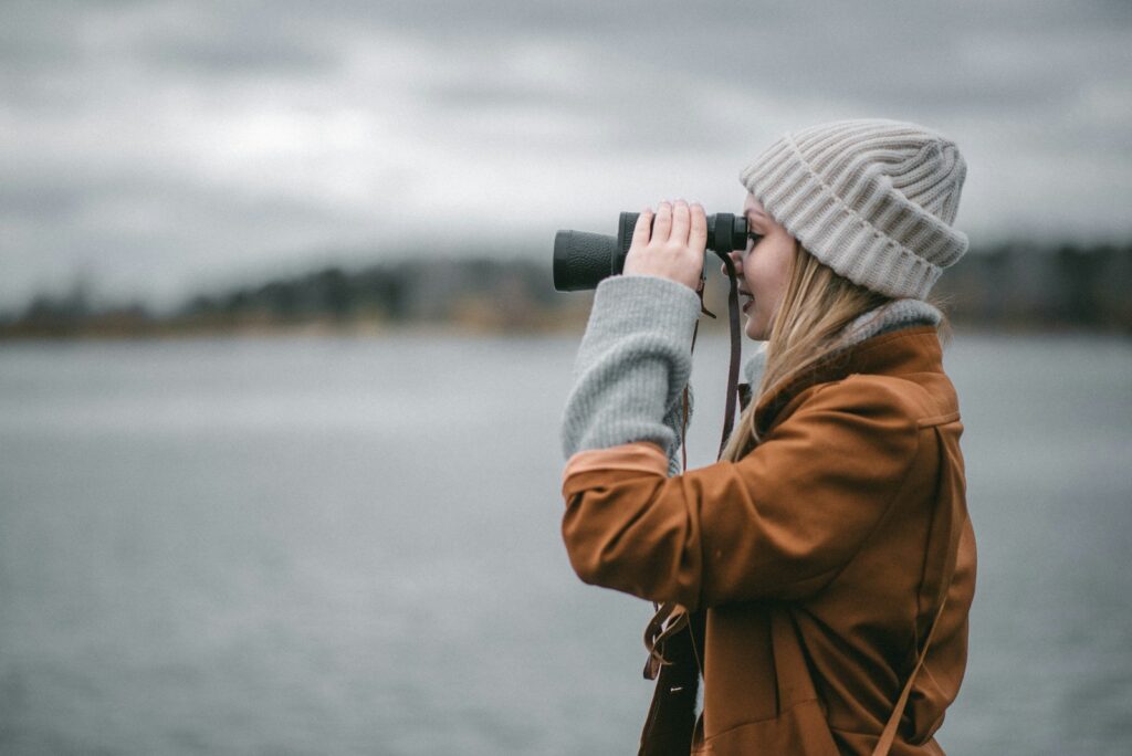 A person in a tan coat and white beanie looks through black binoculars over a body of water.
