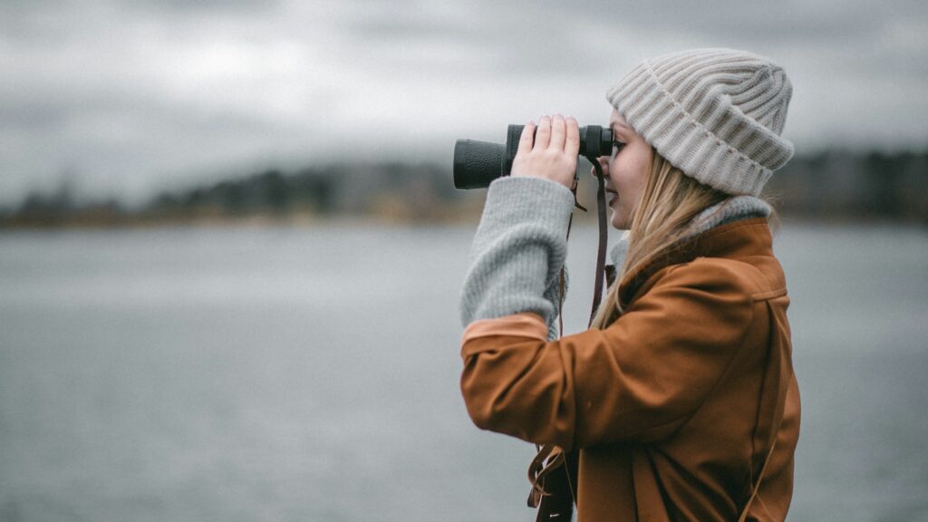 A person in a tan coat and white beanie looks through black binoculars over a body of water.