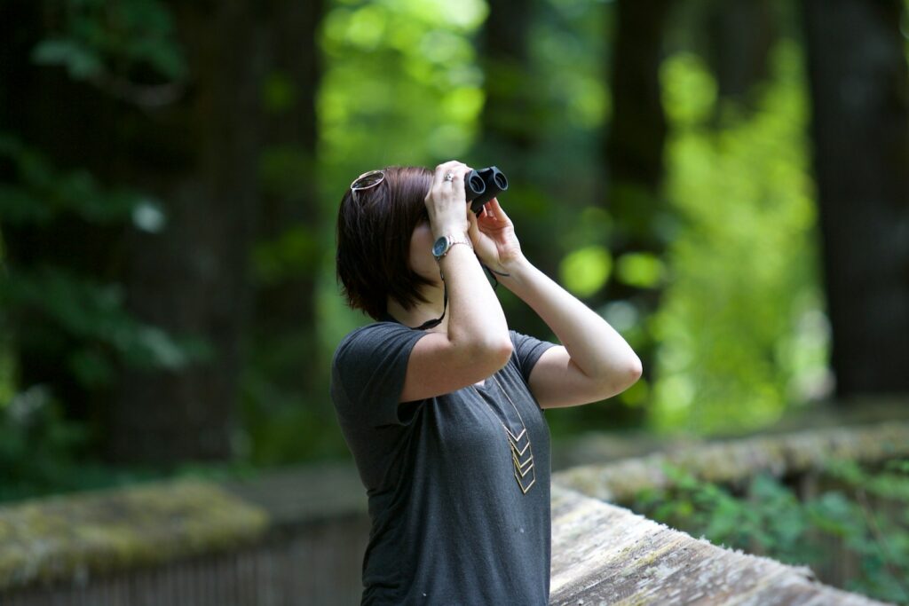 A person with short brown hair uses black binoculars to observe bird in the distance.