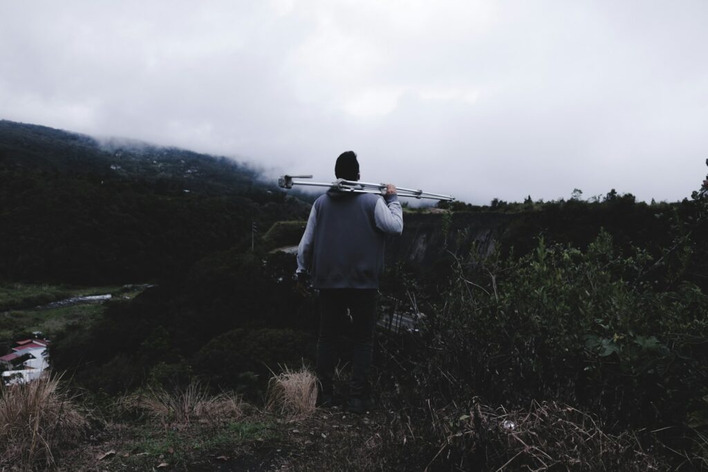 A person carries tripod legs over their shoulder in a hilly, possibly foggy, landscape.