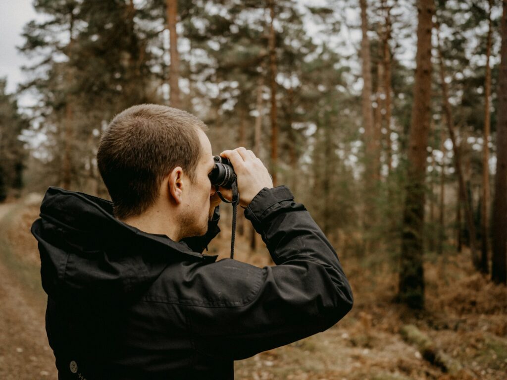 A man in a black jacket uses binoculars while standing among trees.