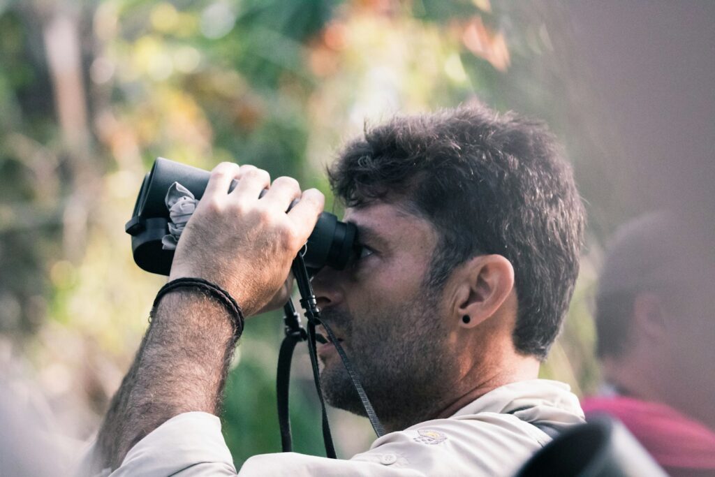 A man with dark hair and a bracelet looks through black binoculars.
