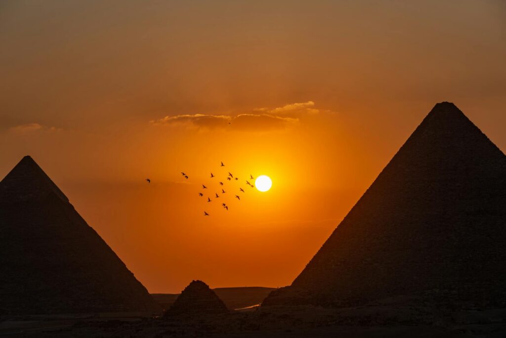 Majestic silhouette of the Giza pyramids at sunset with birds flying against a vibrant sky.