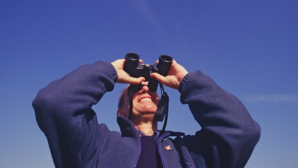 Man birdwatching with binoculars under a clear blue sky