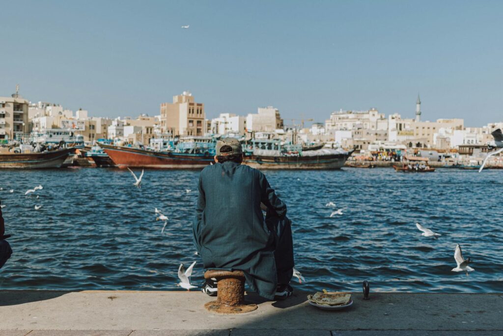 A man sits by the water as several gulls fly around him.
