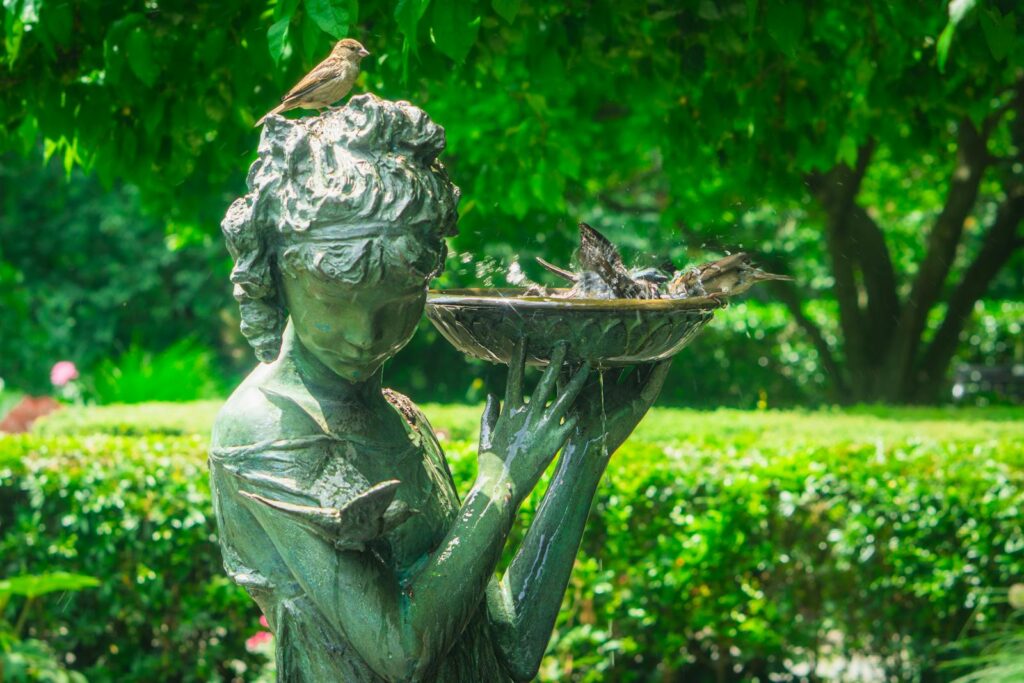 A serene bronze statue in a lush New York garden with birds enjoying a bath under sunlight.