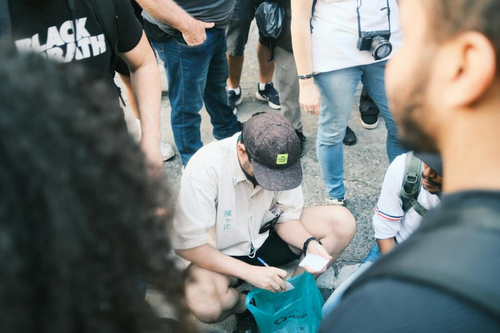 A person in a cap kneels on the ground and writes on a small piece of paper.