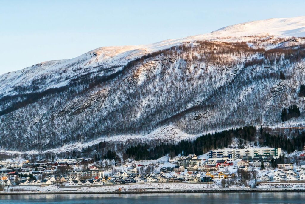 A scenic winter view of a Norwegian town beneath snowy mountains reflecting in a fjord.