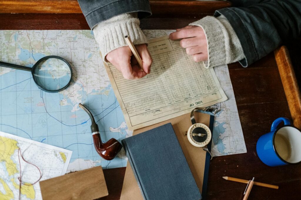 A person writes in a logbook next to a map, a pipe, and a compass.