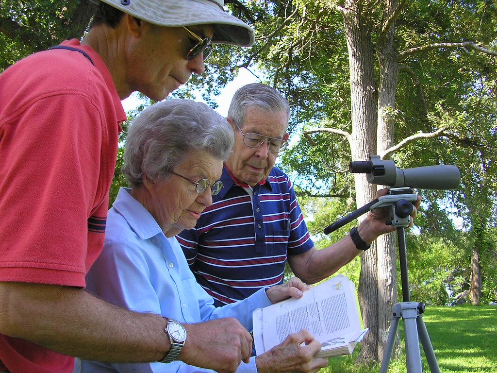 Three individuals are focused on a bird identification book next to a spotting scope.