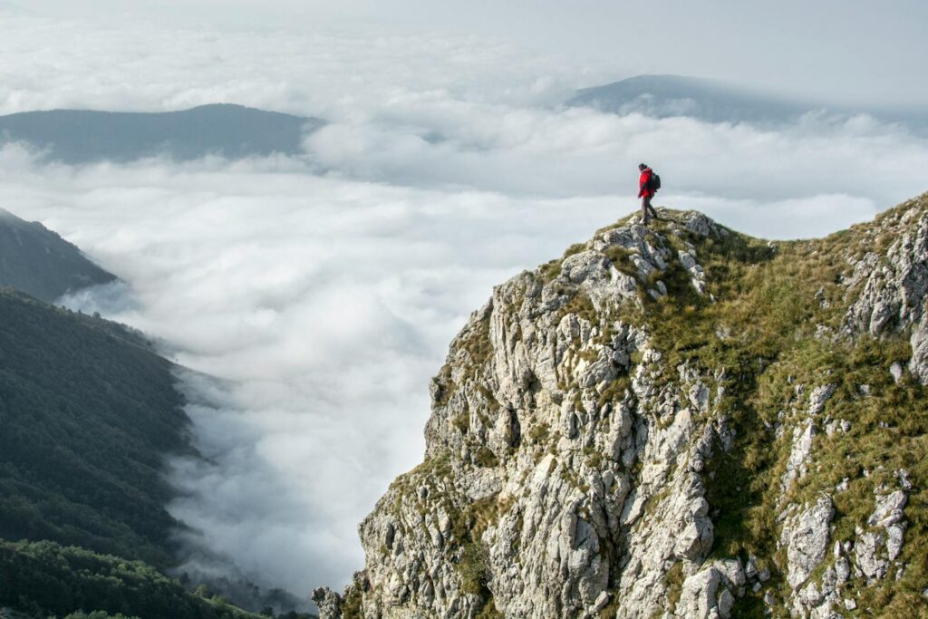 Hiker in red standing on a rocky mountain summit enveloped by clouds.