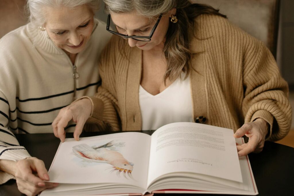 Two elderly women are reading an illustrated book, sharing a moment of connection.