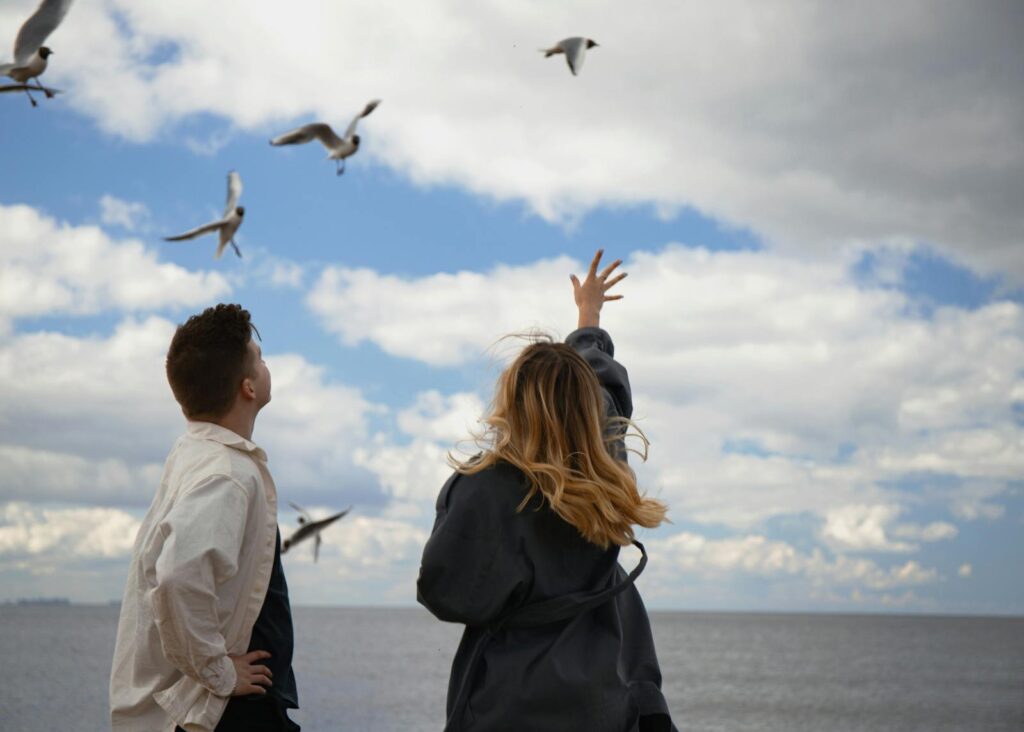 A young couple by the sea watching seagulls in a cloudy sky.