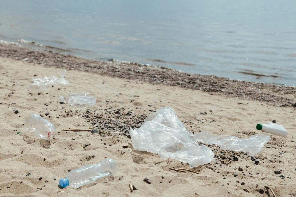 Discarded plastic bottles and bags litter a sandy beach near the water.