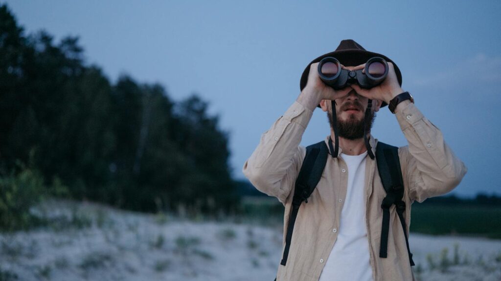 A man birdwatching on a sandy beach wearing a backpack and hat.