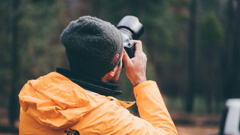 A man in a yellow jacket holding a black DSLR camera in a forest.