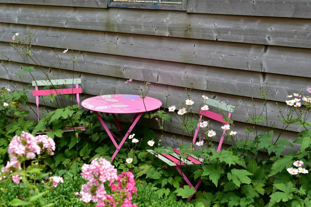 A pink garden table sits among green plants and white flowers.