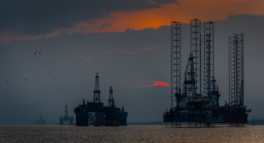 Offshore oil rigs are silhouetted on the water against a dim sky, with birds flying above.