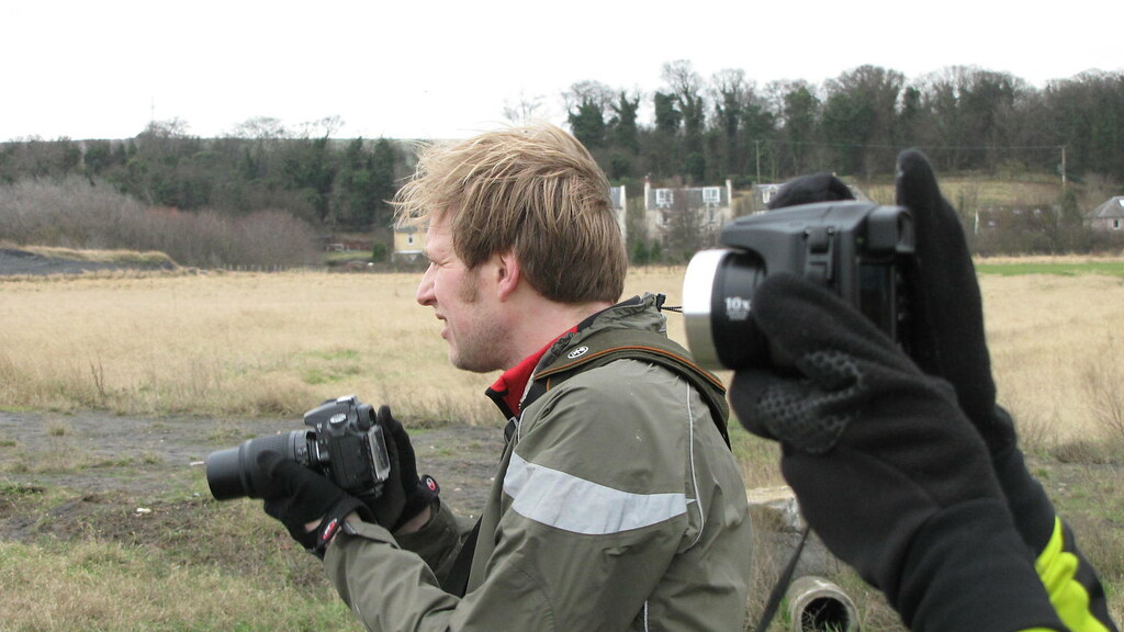 A man with blond hair is holding two cameras outdoors for capturing bird photo.