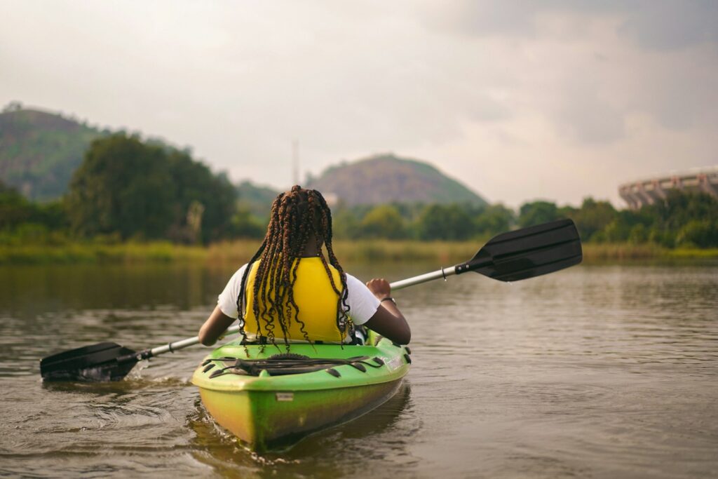 A person in a yellow life vest paddles a green kayak on calm water.