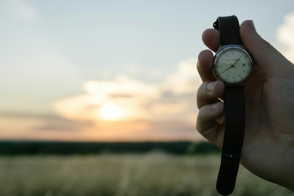 A hand holds a wristwatch against a blurred sky and landscape.
