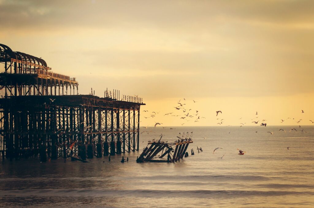 A dilapidated pier stands over choppy water, with many birds in flight under a golden sky.