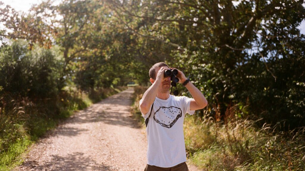 man standing on a dirt road holding binoculars possibly birdwatching