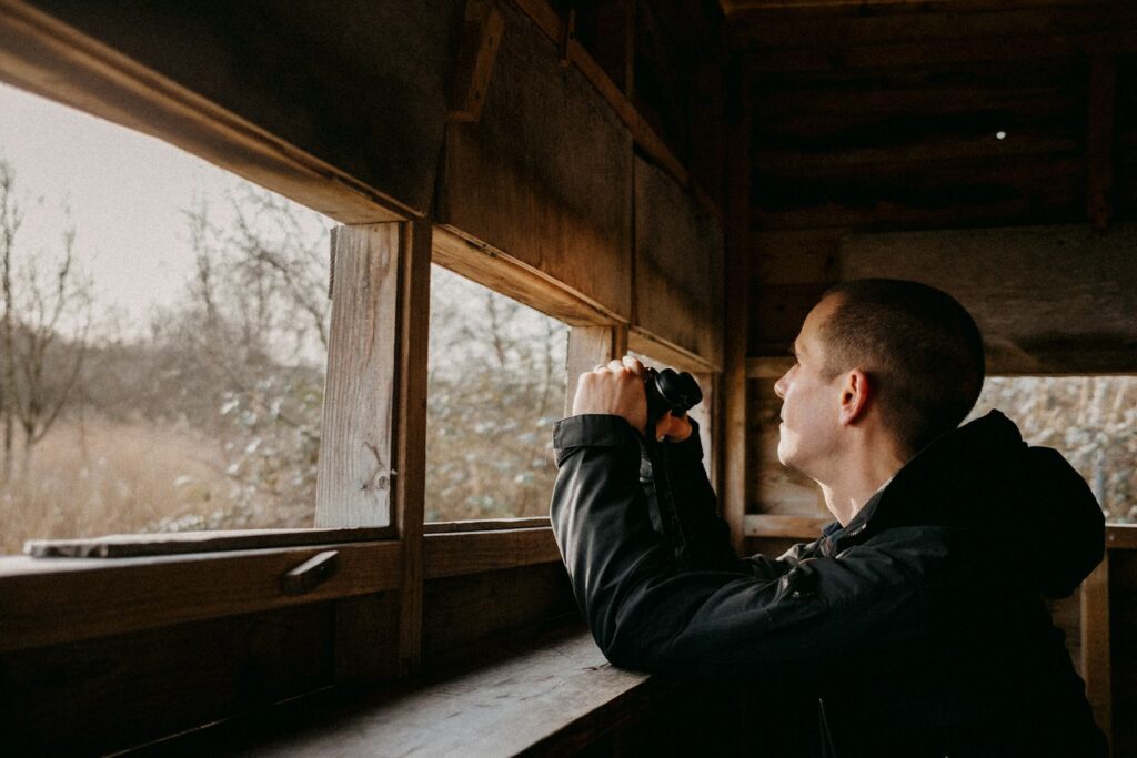 A person uses binoculars to observe birds through a window.