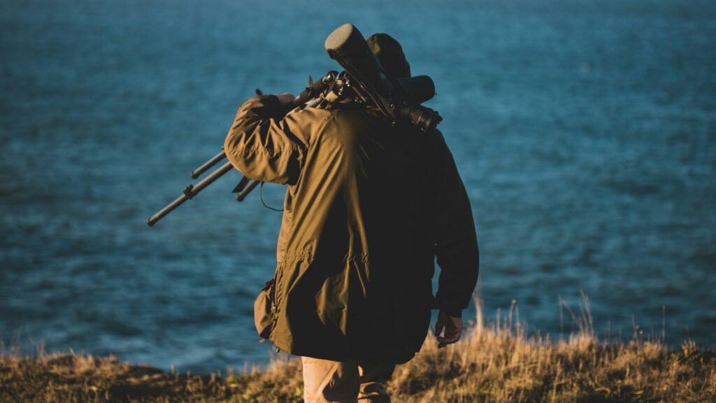 Man in black jacket carrying telescope walking along the ocean shore