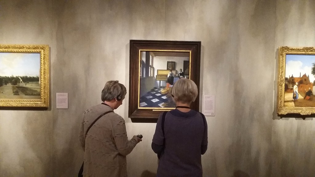 Two people view a framed painting in a gallery setting, with other artworks visible nearby.