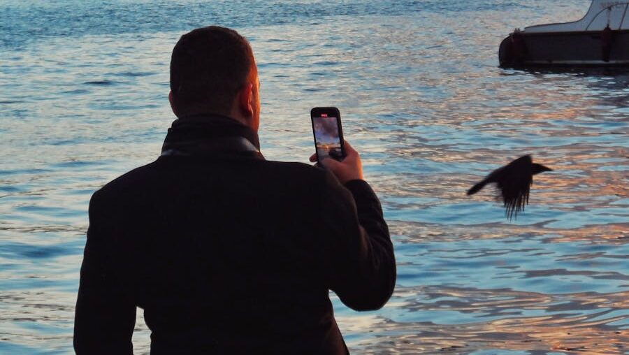 Man photographing bird with smartphone near calm water at sunset