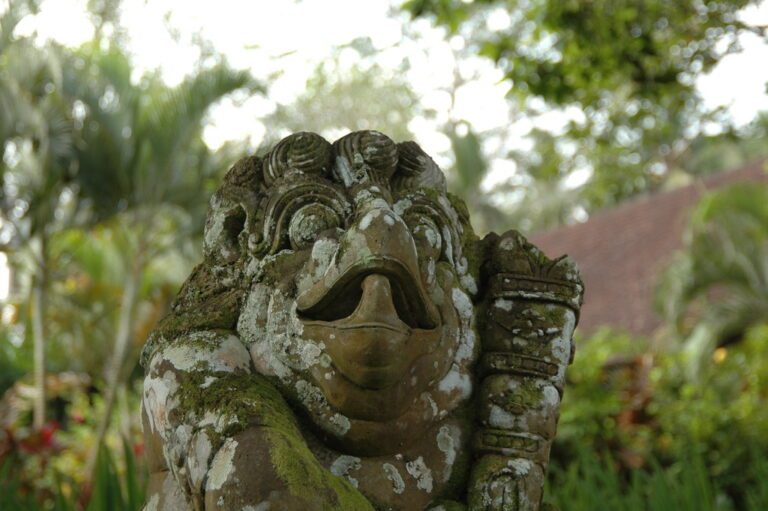 A moss-covered stone statue of a mythical bird with a wide mouth stands amidst lush greenery.