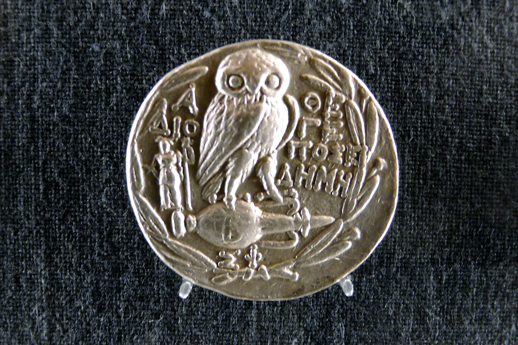 An ancient silver coin depicts an owl with large eyes, surrounded by intricate symbols.