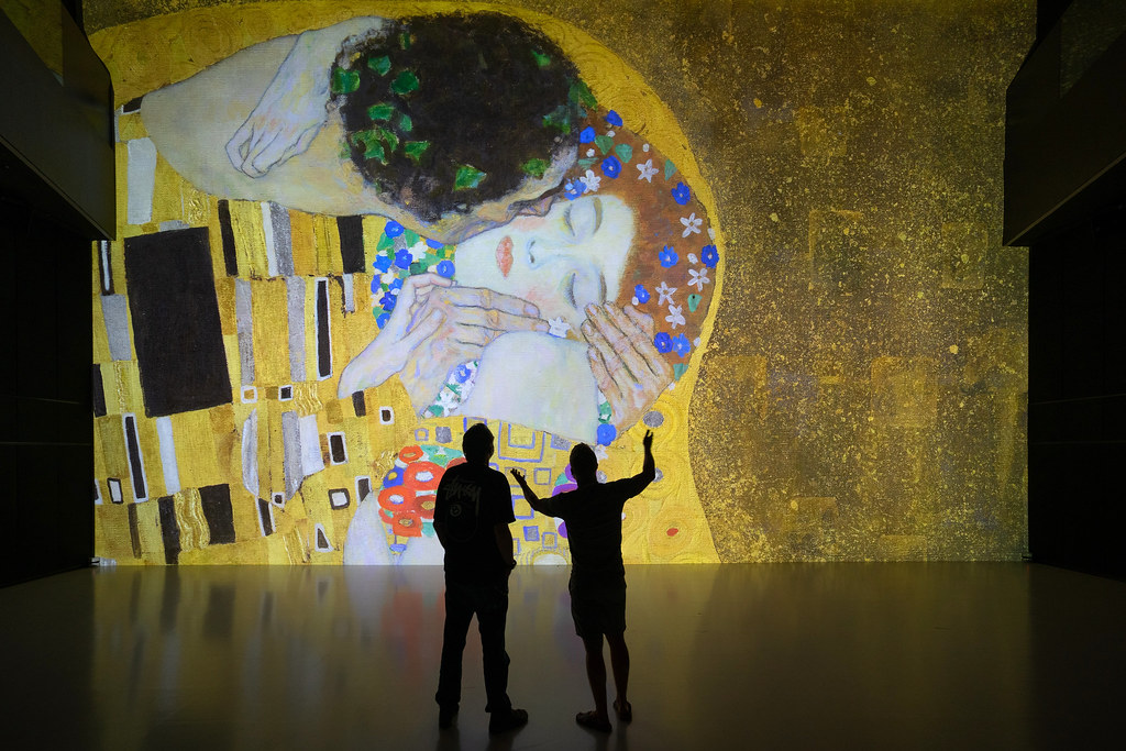 Silhouetted figures stand before a large, illuminated projection of a golden, ornate painting of two embracing people.