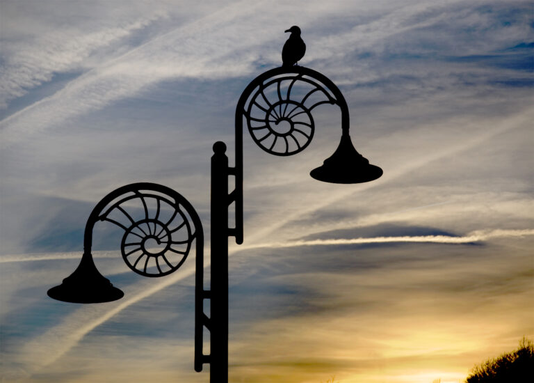 A bird perches atop a decorative, spiral-designed lamp post silhouetted against a cloudy sunset sky.