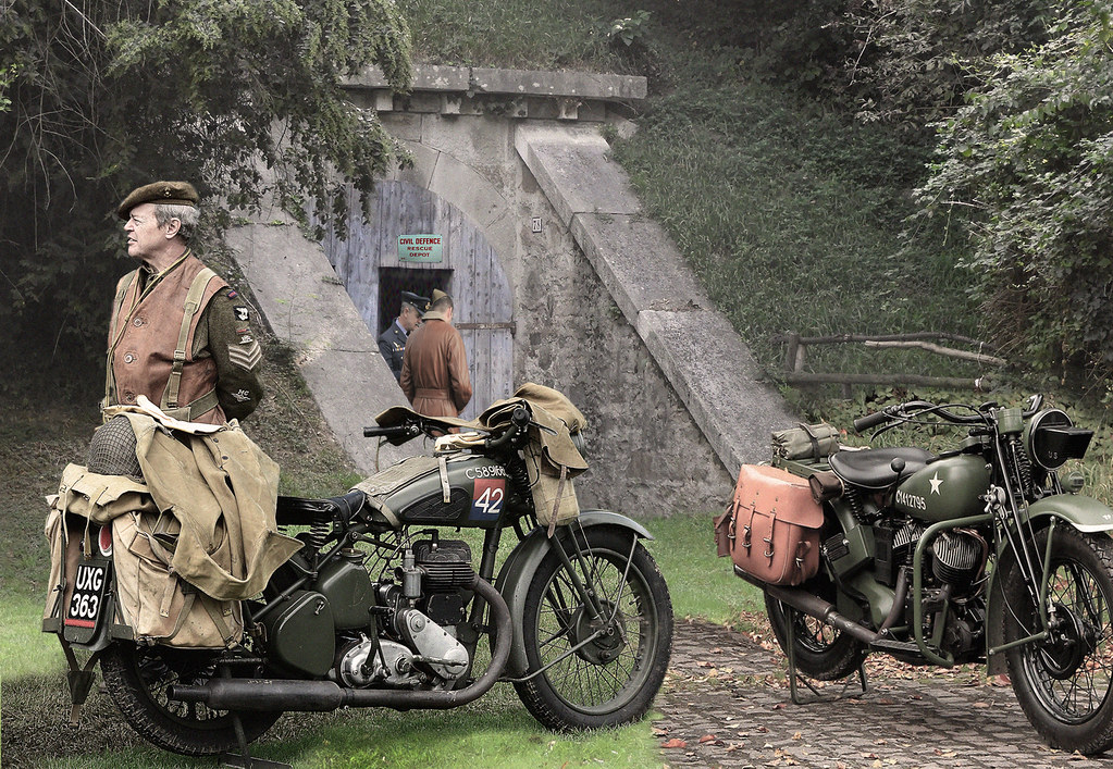 Two vintage military motorcycles are parked near an underground entrance, with a soldier nearby.