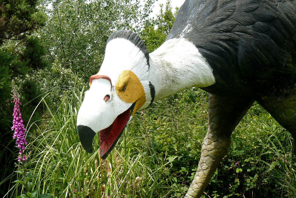 A model of a Phorusrhacos, with a white head, yellow eye patch, and open red beak, among green foliage.